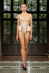Model on a runway wearing a structured, pale pink corset-style bodysuit with cutouts and sheer panels; black heels, dark doorway backdrop.