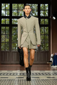 Runway look: oversized beige blazer with lace-up side detailing, matching shorts, and black platform boots under bright show lights.