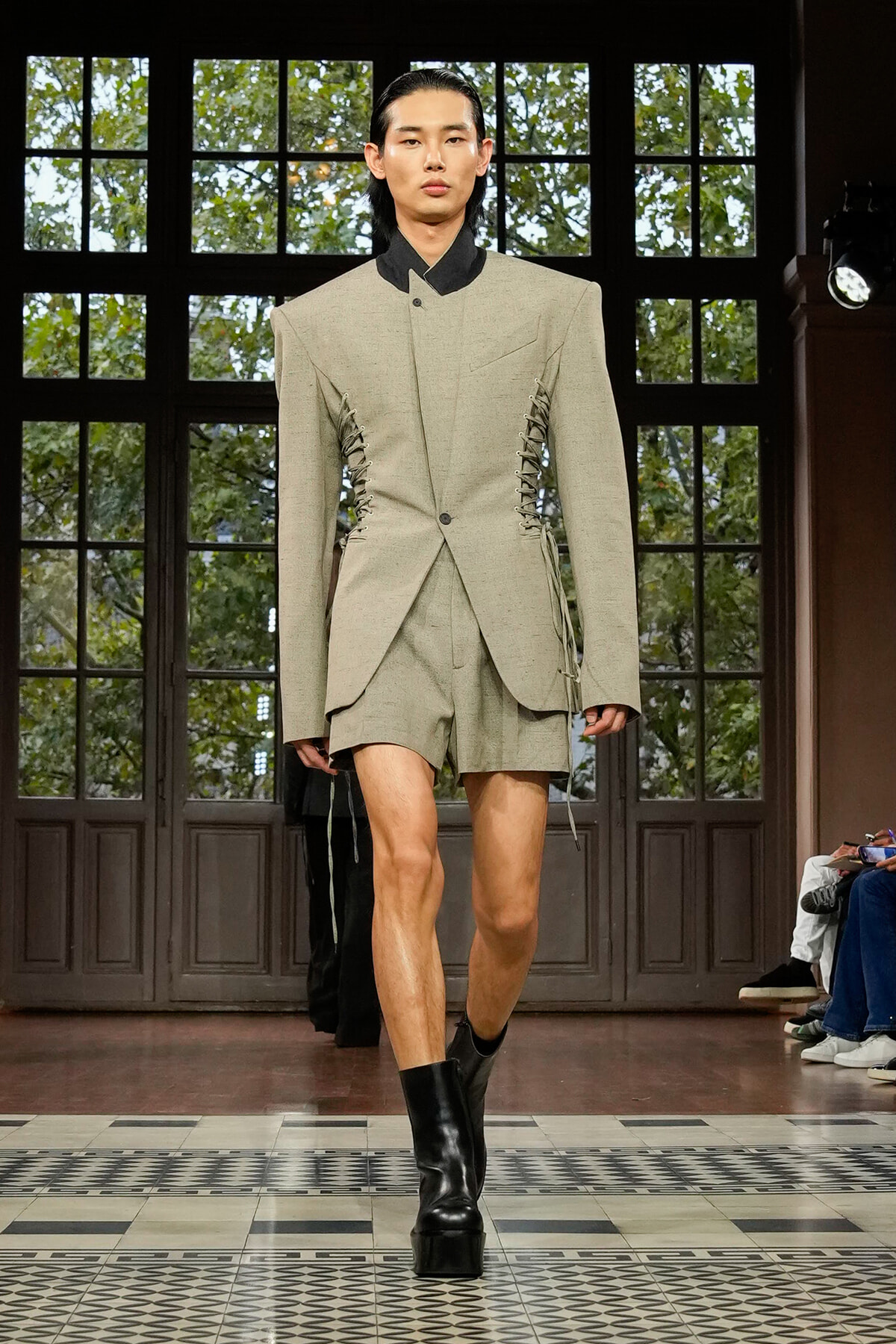 Runway look: oversized beige blazer with lace-up side detailing, matching shorts, and black platform boots under bright show lights.