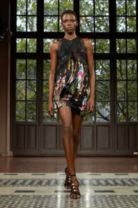 Runway model wearing a black, paint-splattered dress with sheer top and strappy pearl-embellished heels walking down a tiled corridor runway.