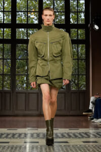 Runway model wearing an olive green utility jacket with oversized chest pockets and matching shorts, mid-stride on a fashion show runway