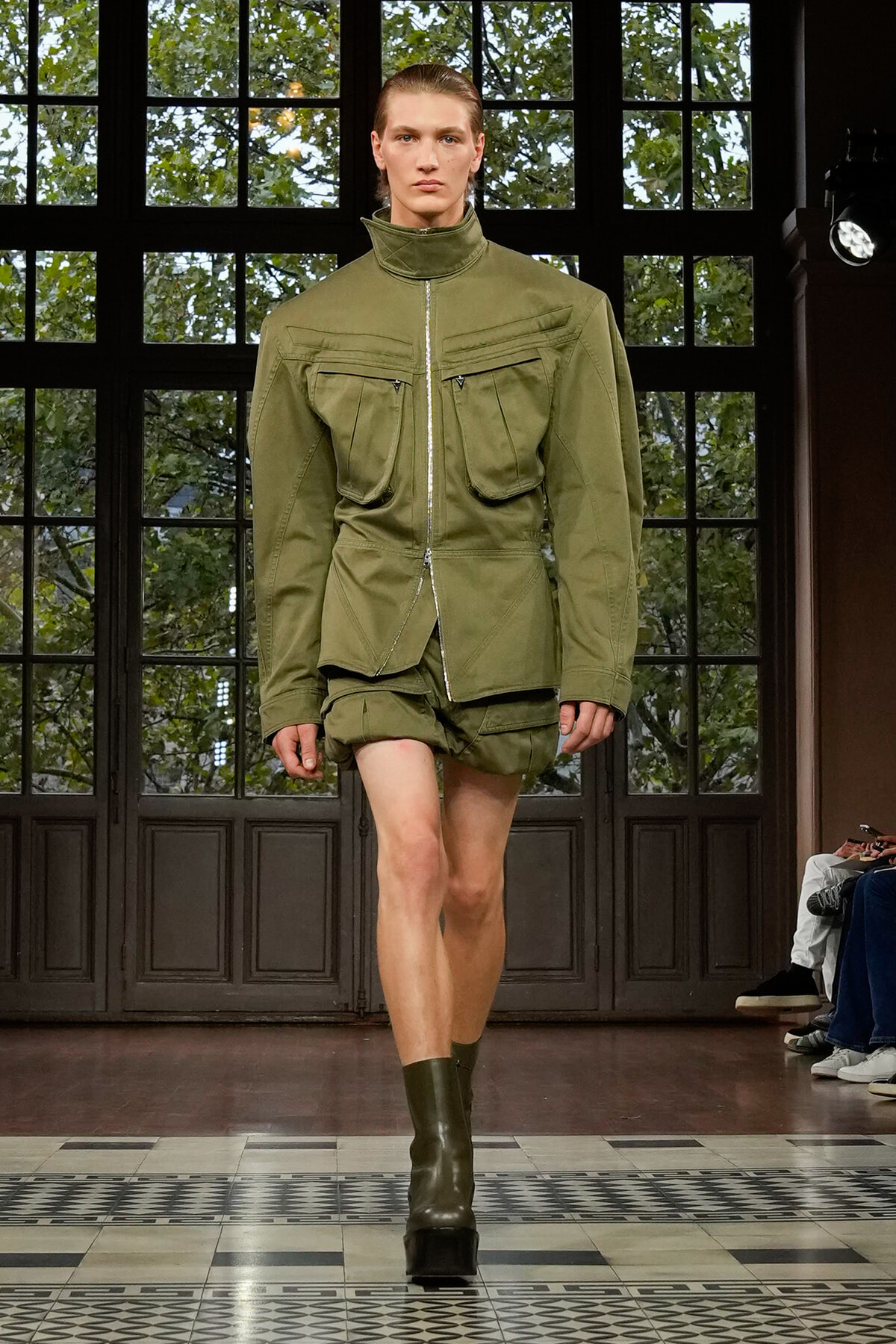 Runway model wearing an olive green utility jacket with oversized chest pockets and matching shorts, mid-stride on a fashion show runway