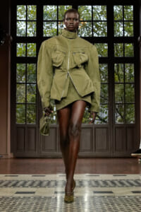 Model walks the runway in an olive-green oversized utility jacket with a high collar, zippered pockets, and a short matching skirt, carrying a small olive bag.