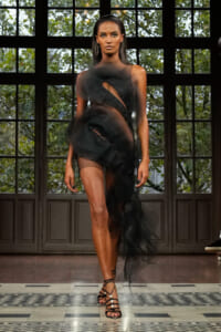 Model walking a runway in a sheer black cutout gown with strappy heels, against large grid-window backdrop.