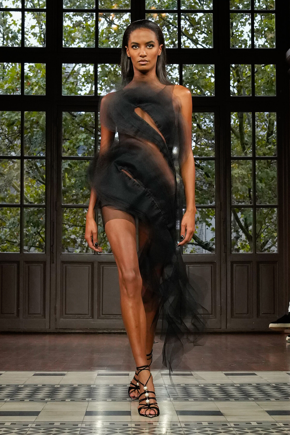 Model walking a runway in a sheer black cutout gown with strappy heels, against large grid-window backdrop.