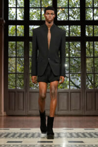 Male model on a runway wearing a black blazer with no shirt, matching black shorts, and black ankle boots.