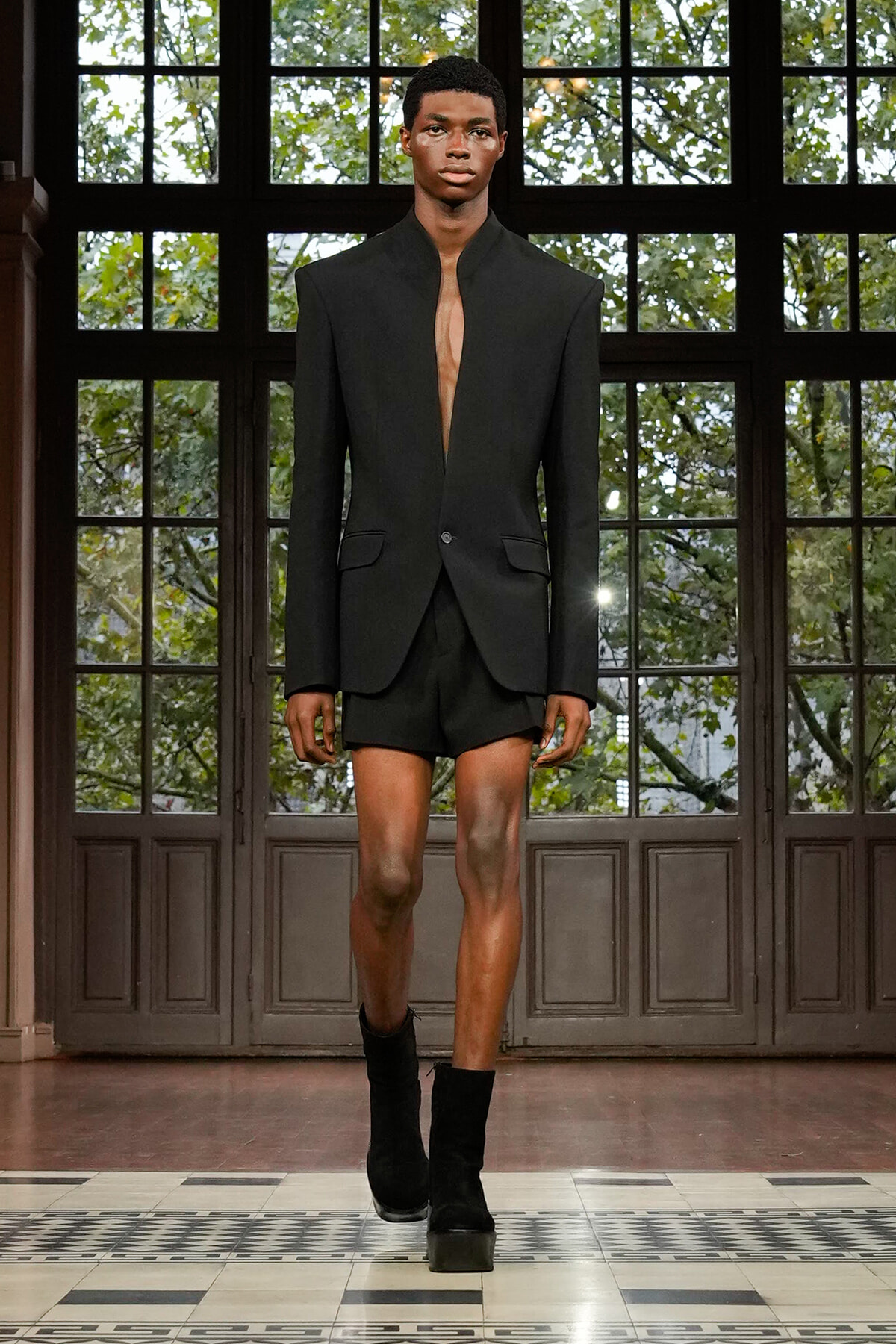 Male model on a runway wearing a black blazer with no shirt, matching black shorts, and black ankle boots.