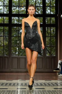 Model on runway wearing a black sequined strapless dress with a deep V and asymmetric mini hem, paired with black ankle-strap heels.