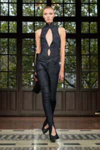 Model walks the runway in a dark denim jumpsuit with a high collar and cutouts, carrying a black clutch.