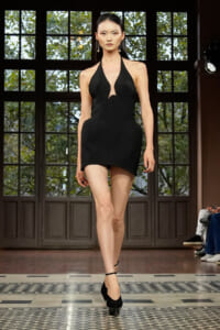 Model walking a runway in a black halter-neck mini dress with a plunging neckline and sculpted panels, indoors by large windows.