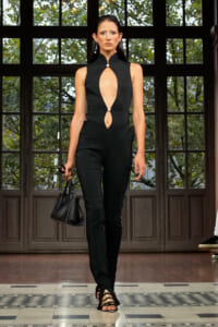 Model walking runway in a black sleeveless jumpsuit with large chest and midsection cutouts, pearl buttons, and a black handbag.