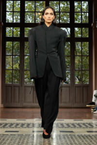 Model on a runway wearing a dramatic black tailored blazer with corset-style side lacing and wide shoulders, paired with loose black pants.