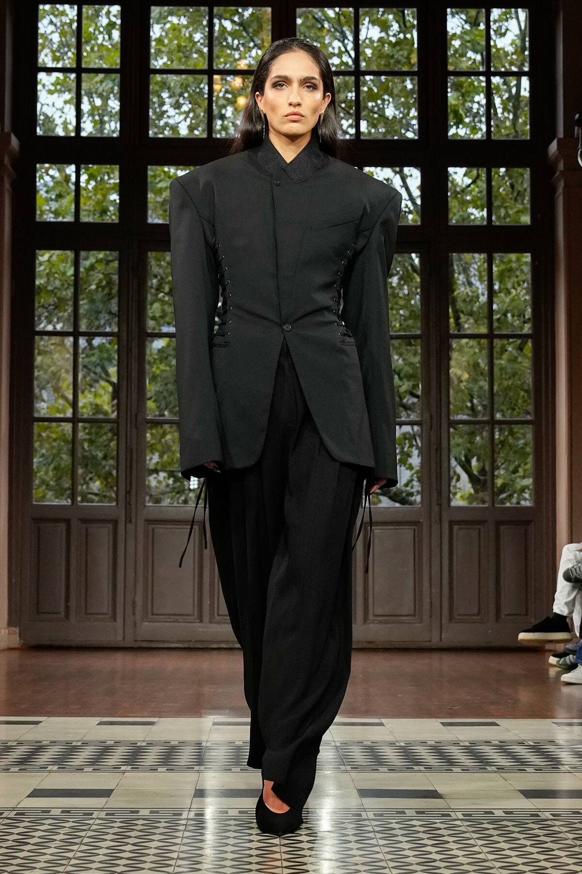 Model on a runway wearing a dramatic black tailored blazer with corset-style side lacing and wide shoulders, paired with loose black pants.