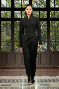 Model walks the runway in a fitted black sculptural blazer and matching trousers with strappy sandals.
