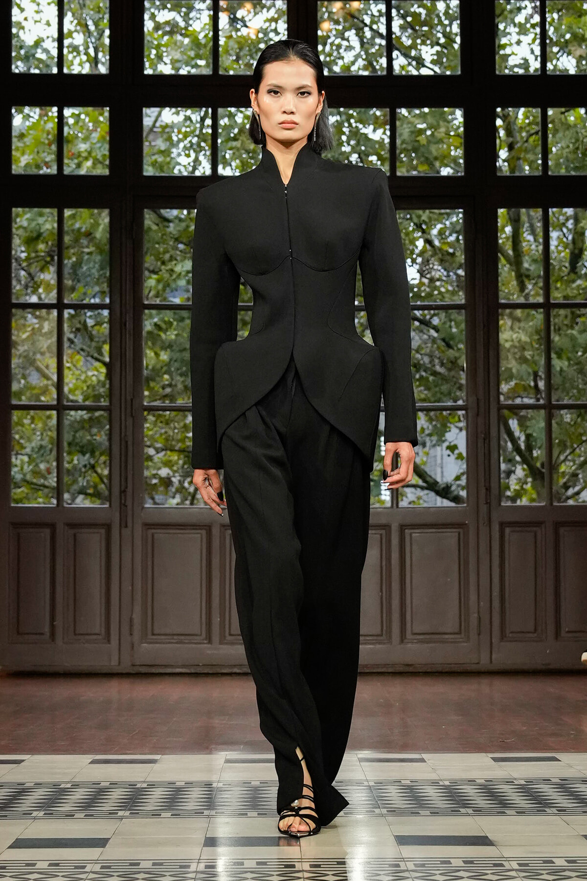 Model walks the runway in a fitted black sculptural blazer and matching trousers with strappy sandals.