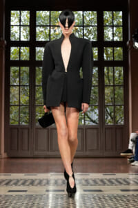 Model walks a runway in a structured black blazer dress with a deep plunging neckline, pairing it with black heels and a small black handbag in a gallery-style venue.
