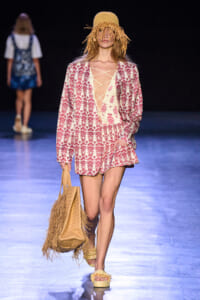 Model walking a runway in a pink-and-cream patterned blazer/romper with a lace-up front, straw hat with fringe, and a fringed straw tote bag. Behind her, another model is blurred.