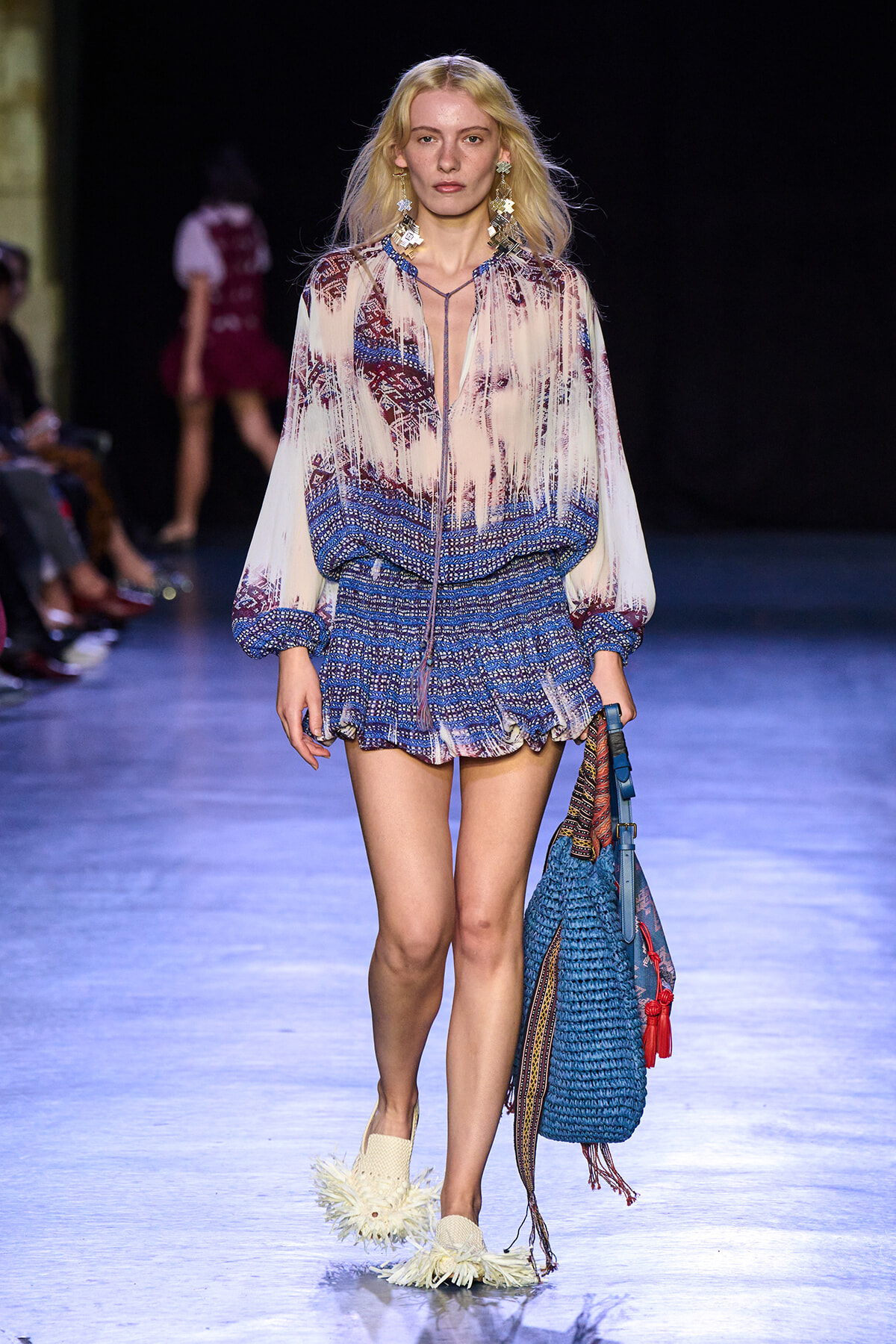 Model on a runway wearing a sheer printed blouse with blue patterned shorts and a large blue woven bag on her side.