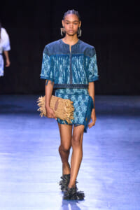 Model on a runway wearing a blue denim-patterned two-piece with fringe sleeves, holding a woven straw clutch at the hip.