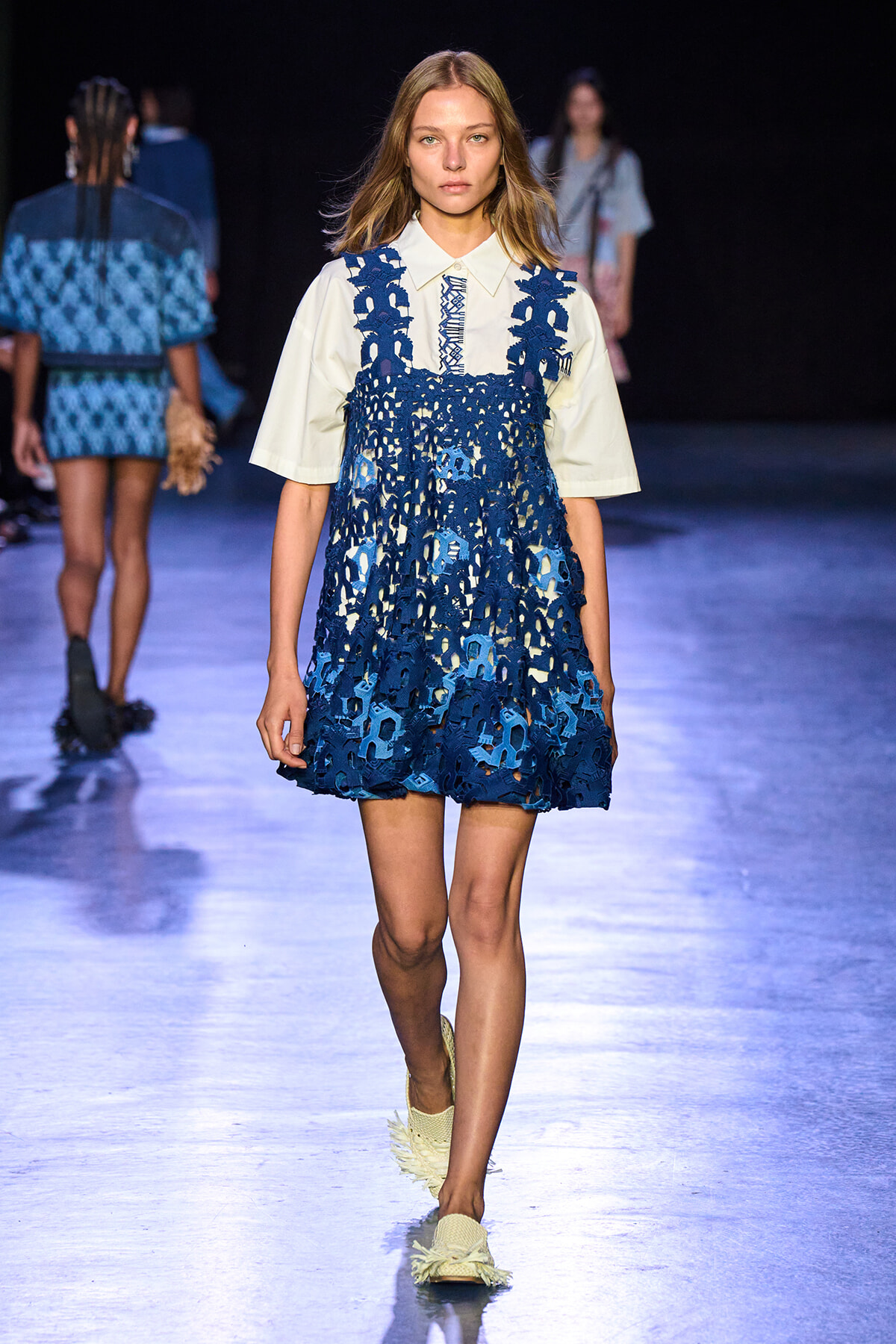 Model walking the runway in a white collared shirt layered under a blue crochet overlay dress with circular patterns, on a fashion show catwalk.