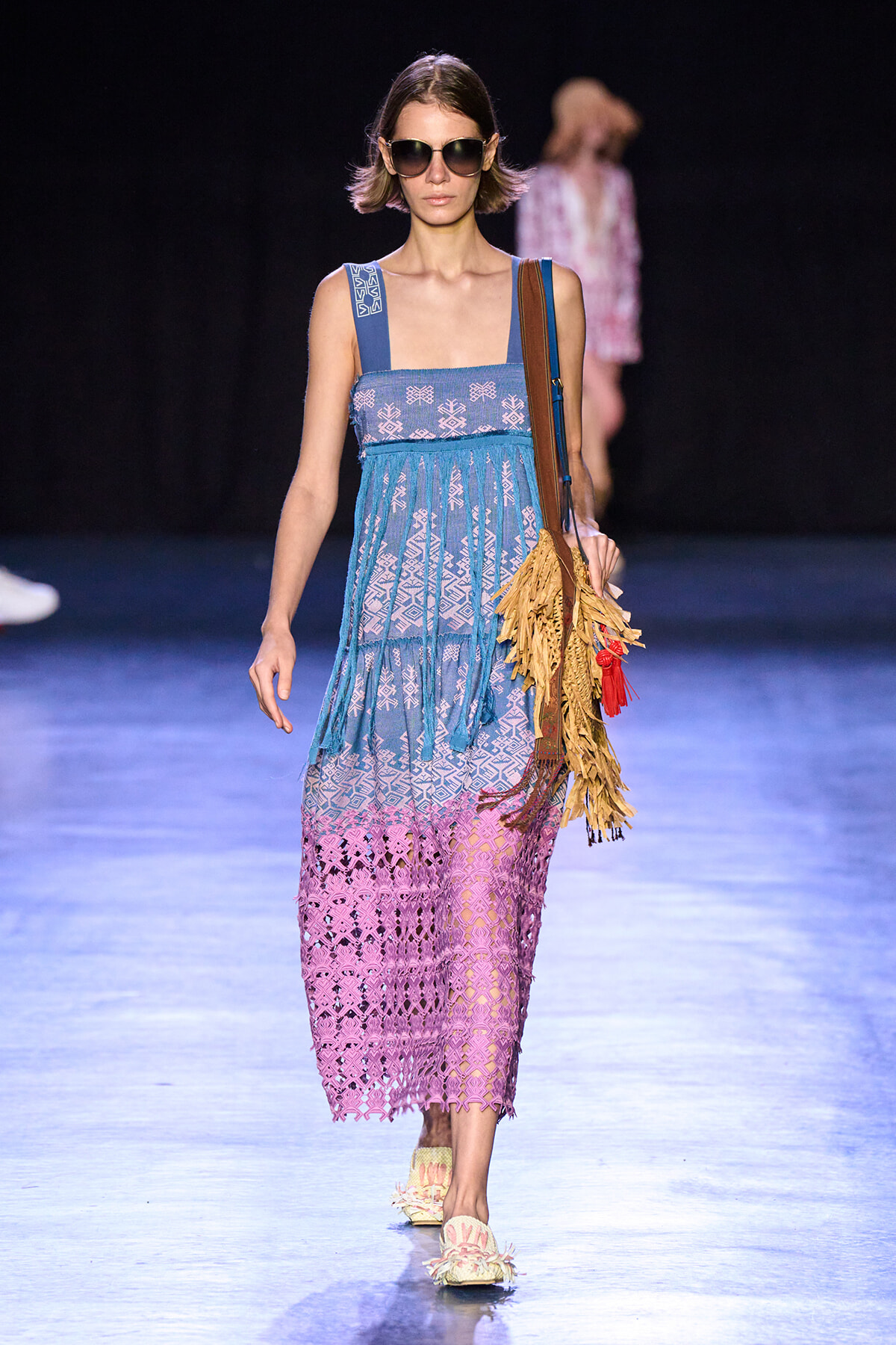Runway model wearing a blue‑to‑pink crocheted maxi dress with geometric patterns, oversized sunglasses, and a fringe tan tote bag.