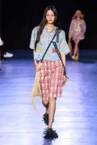 Model walks the runway in a light blue top with fringe patches, red geometric shorts, a woven fringe bag, and spiky black footwear.