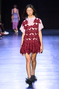 Model walking runway in a burgundy lace overlay dress over a pale pink blouse, paired with feathered black shoes and glasses.