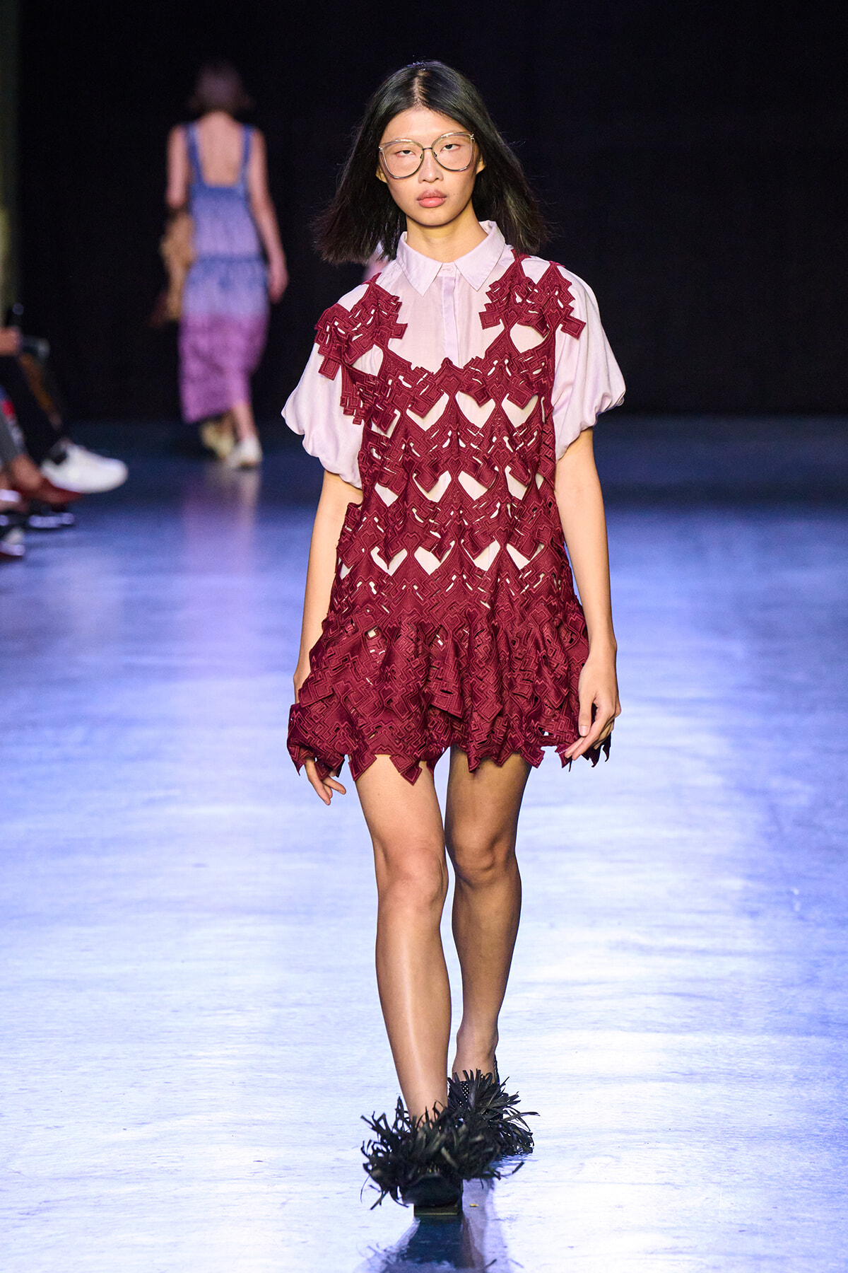 Model walking runway in a burgundy lace overlay dress over a pale pink blouse, paired with feathered black shoes and glasses.