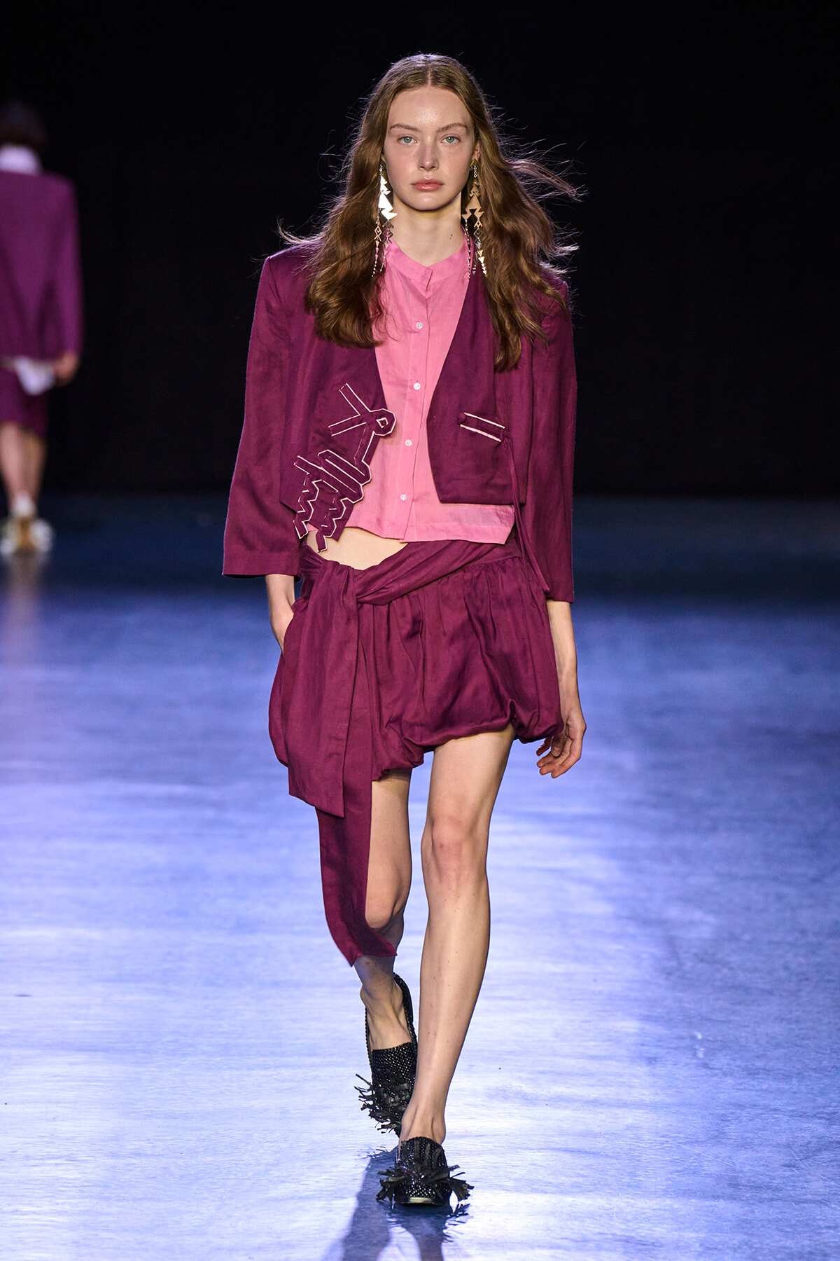 Model walking the runway in a pink shirt paired with a magenta jacket and draped shorts, accessorized with long geometric earrings.