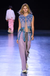 Model walking the runway in a sleeveless blue patterned top with long turquoise fringe, lavender wide-leg pants, and a beaded belt.