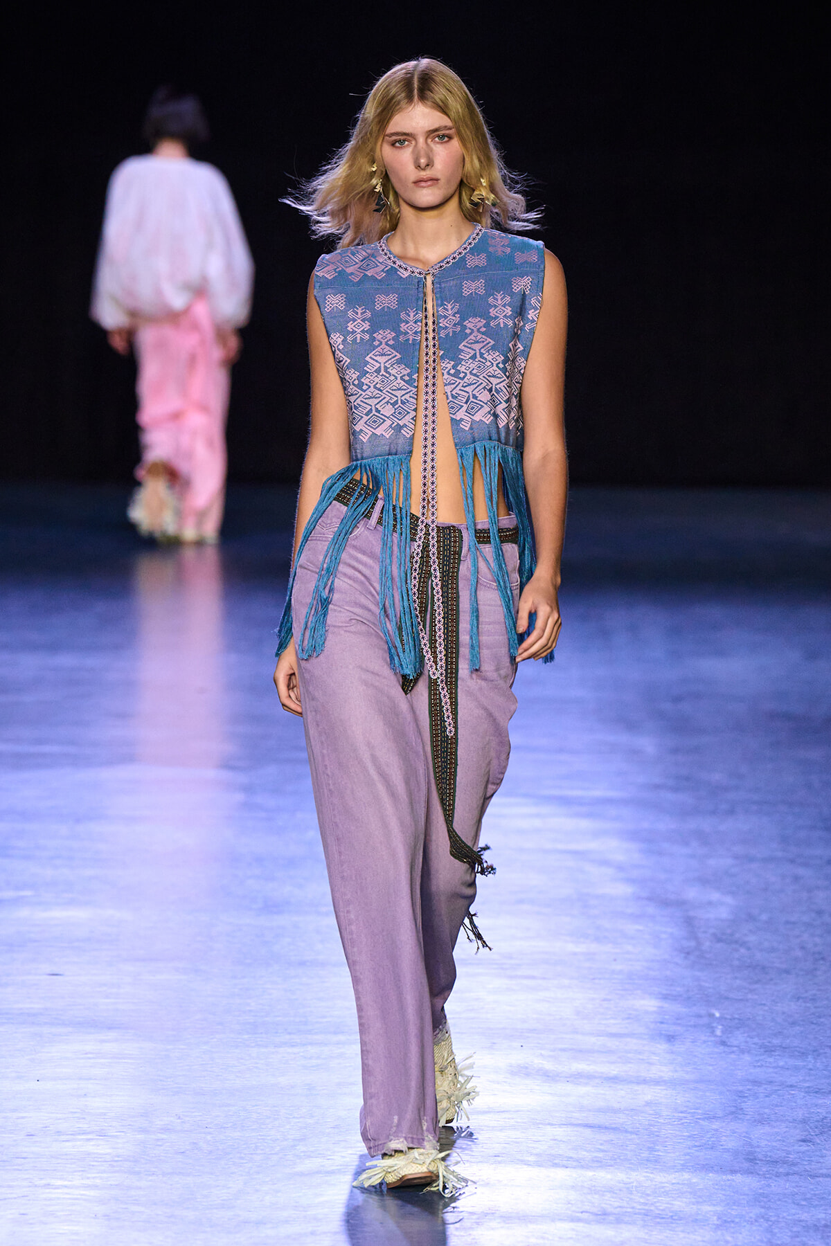 Model walking the runway in a sleeveless blue patterned top with long turquoise fringe, lavender wide-leg pants, and a beaded belt.