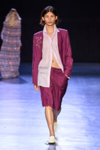 Model on a runway wearing a magenta suit with a light pink shirt, hand in pocket, spotlighted on dark stage.