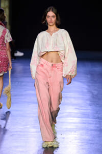 Model walks the runway in a cropped white blouse with pink print and high-waisted pink wide-leg jeans, wearing feathered shoes.