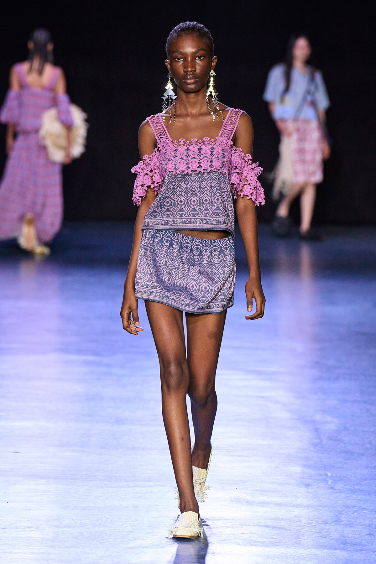 Model walking runway in pink crocheted cold-shoulder top and matching patterned shorts, large geometric earrings, on a blue-lit catwalk