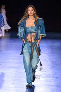 Model walking a fashion show in a blue denim ensemble: cropped fringe-top with fringe strands, matching denim jacket draped over shoulders, and wide-leg jeans with a patterned belt and extra tassels, on a glossy runway.