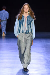 Model walks the runway in a light blue blouse with a dark blue embroidered fringe vest and loose gray trousers, wearing large geometric earrings.