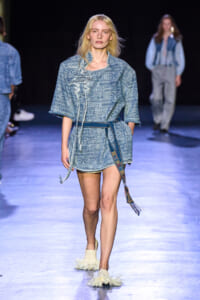 Model walks the runway in a light blue, patterned denim tunic with a belt and fringed slippers.