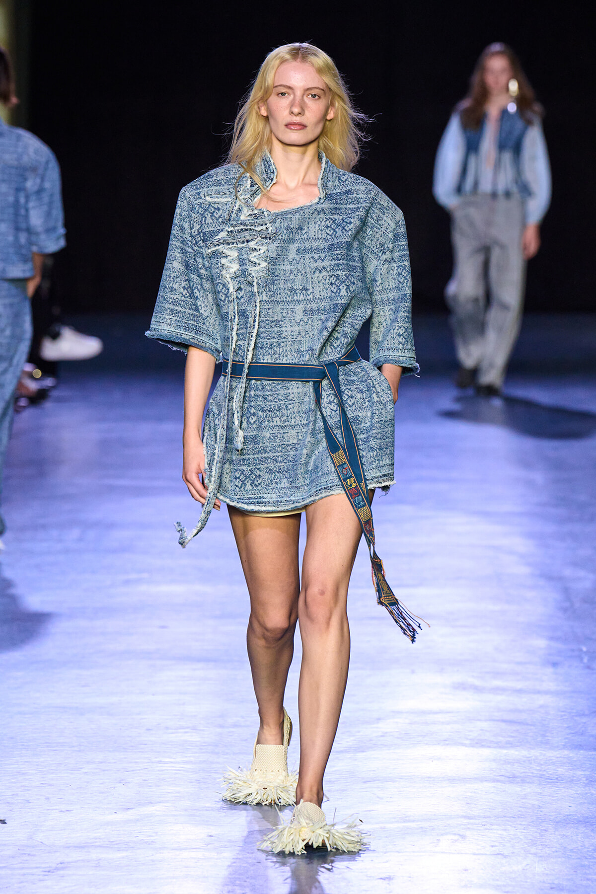 Model walks the runway in a light blue, patterned denim tunic with a belt and fringed slippers.
