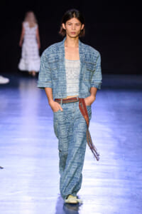 Female model in a blue patterned denim suit walking a runway with a white top and brown tassel belt.
