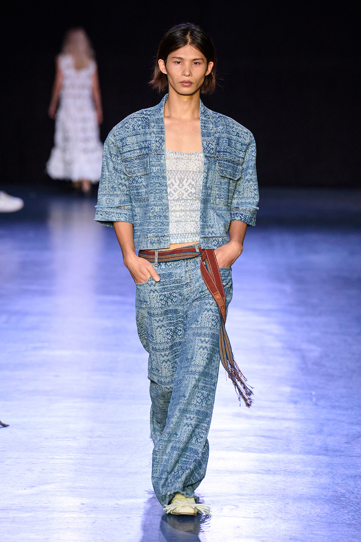 Female model in a blue patterned denim suit walking a runway with a white top and brown tassel belt.