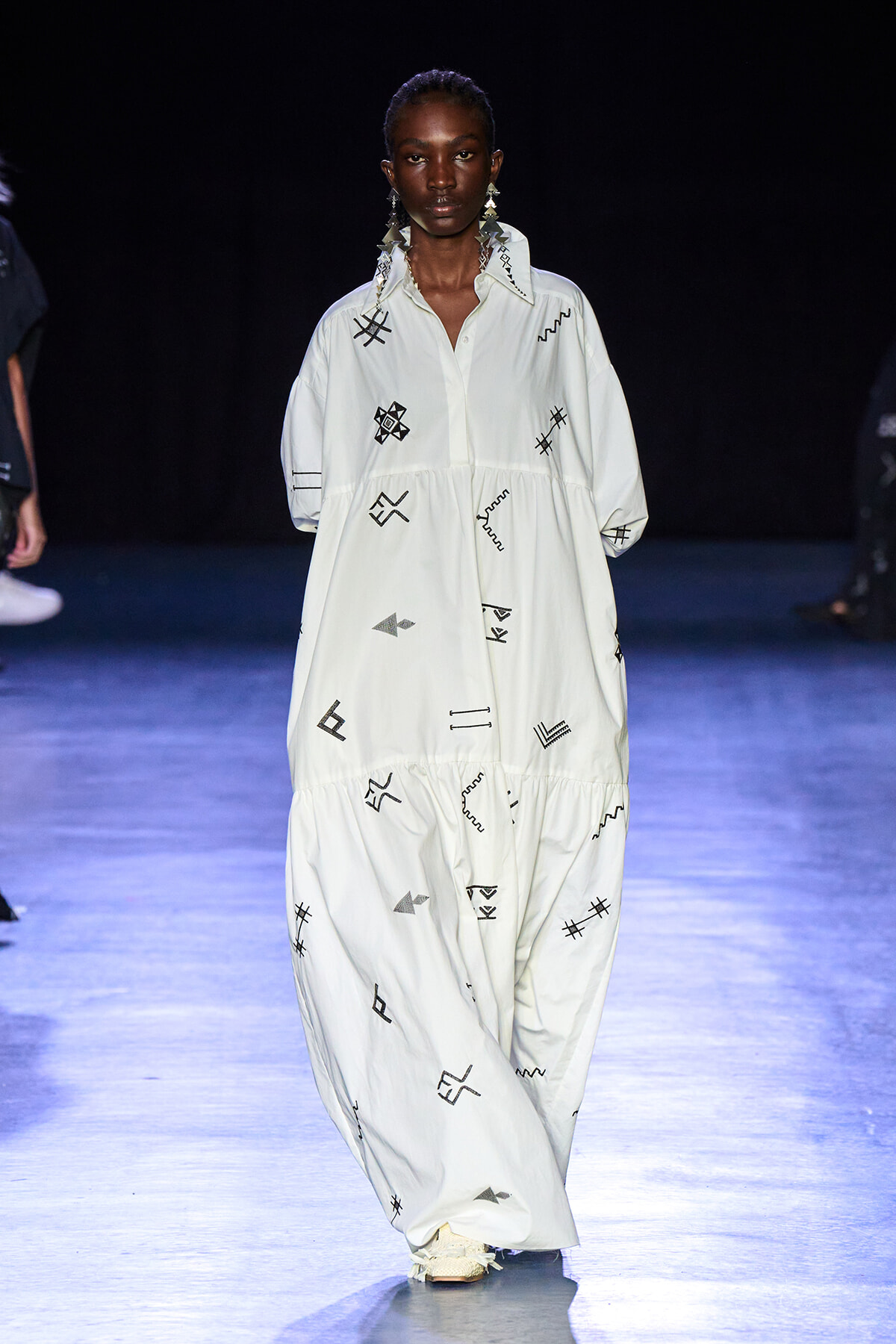 Female model walking a runway in a loose white maxi dress with black abstract symbols.