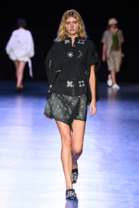 Female model walks a fashion runway in a black oversized shirt with white geometric embroidery and a gray wrap skirt on a blue-lit catwalk.