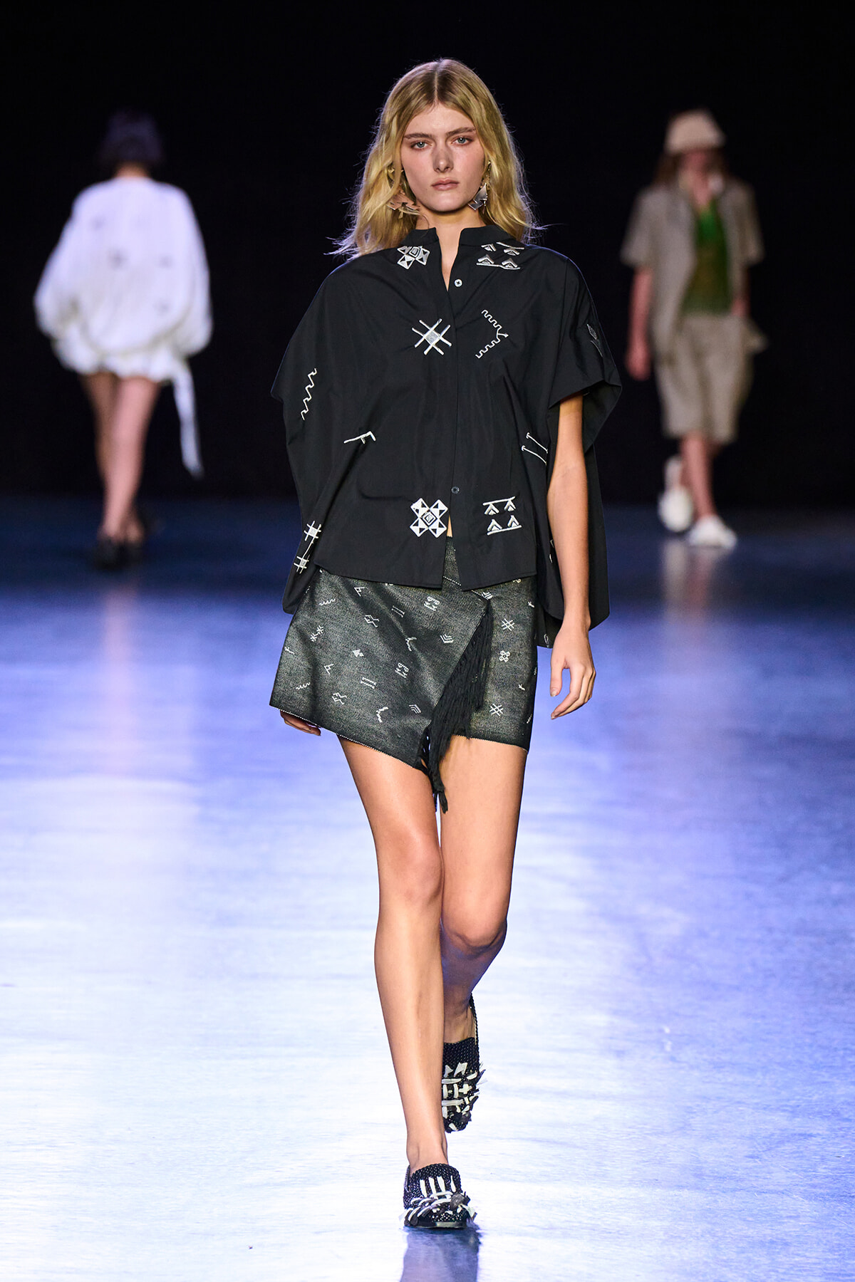 Female model walks a fashion runway in a black oversized shirt with white geometric embroidery and a gray wrap skirt on a blue-lit catwalk.