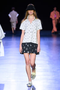 Model walking a fashion runway in a white short-sleeve shirt with black print and a black two-tone skirt, wearing a black visor and woven slides.