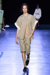 Model walking the runway in a beige cape-style jacket with white cuffs and matching shorts; geometric embroidery on the shoulder and hands in pockets.