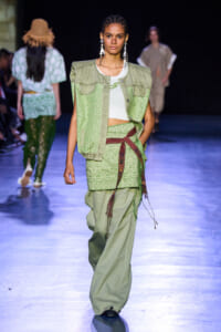 Model walking a fashion show wearing a light-green patterned sleeveless jacket over a white crop top and matching patterned pants with a brown belt to the side of her hip.