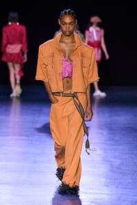 Model walking a fashion runway in an orange cropped jacket and matching wide-leg pants, pink lace top peeking out, with a patterned belt and black spiked boots behind.