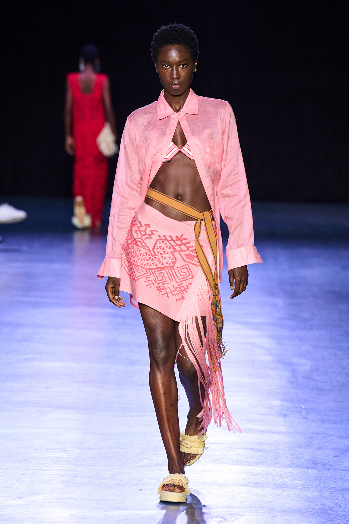 Model walking a runway in a pink sheer shirt over a pink bikini top and a geometric-print fringe wrap skirt with a tan belt.