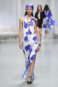 Model walking a runway in a white halter maxi dress with bold blue‑purple flowers and a matching headscarf, black sandals visible.