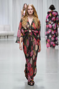 Model walks a runway in a sheer black floral gown with a waist knot and circular cutout at the midriff.